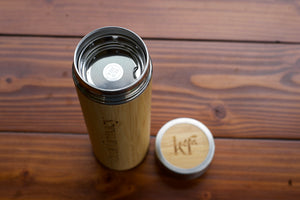 A bamboo insulated travel tumbler with a stainless steel lid and tea infuser, shown next to its bamboo lid on a dark wood plank surface.