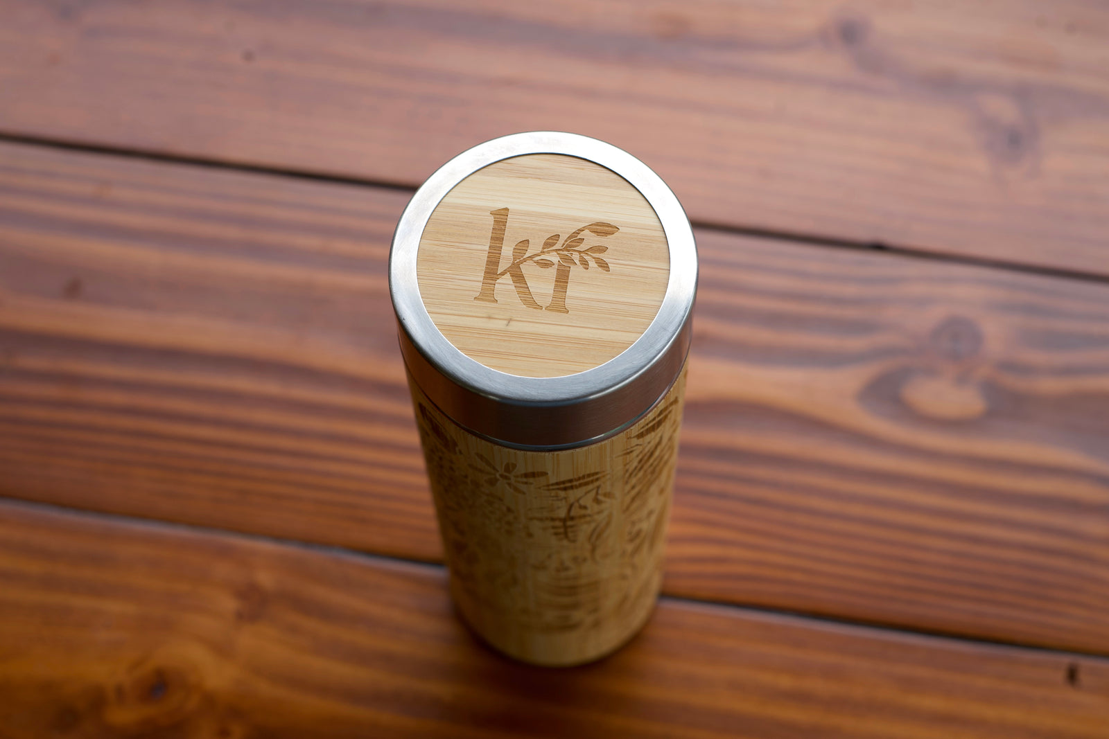 Two bamboo travel tumbler bottles with stainless steel lids sit on a wooden table outdoors, with a green leafy garden in the background. One tumbler is plain with the brand name 'Kaulai Pharmacy' engraved vertically, and the other has an engraved botanical pattern.